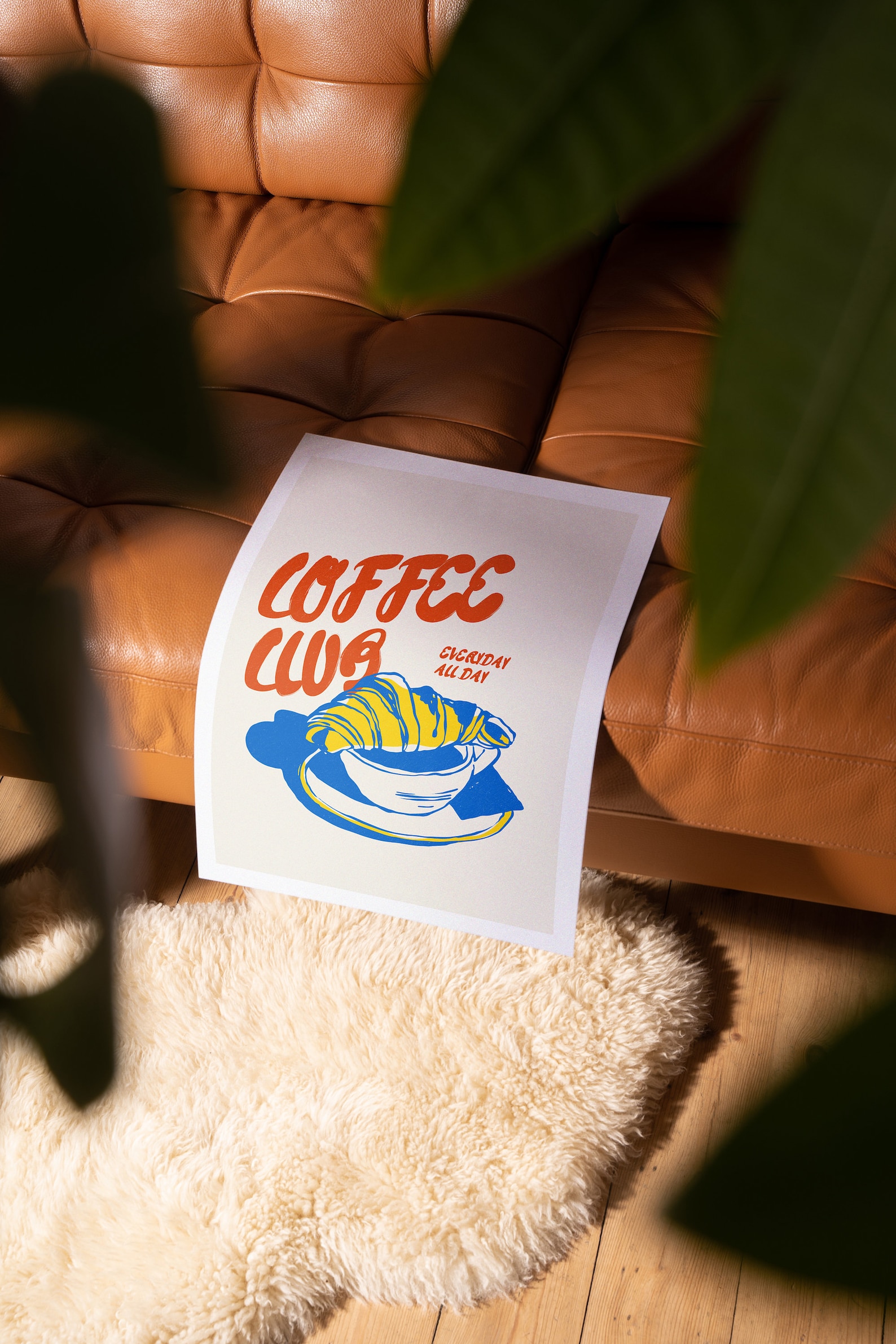 Free poster mockup: Vibrant poster leans on tan leather couch amid sunlight-filtered greenery, on a plush cream rug.